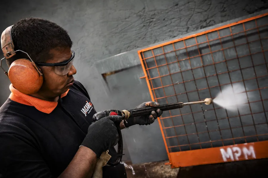 Técnico da MPM lavando com jato de alta pressão uma grade de proteção laranja, na oficina.