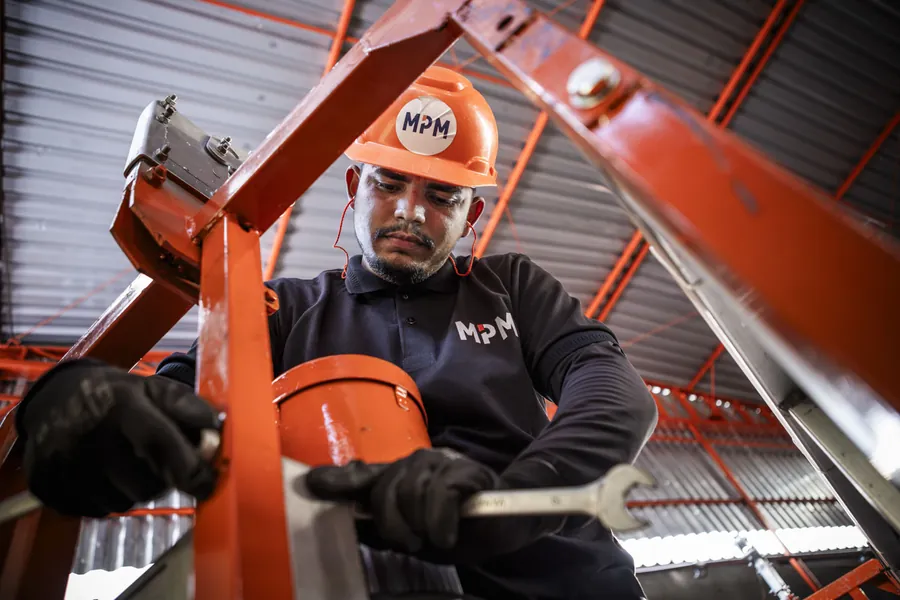 Técnico da MPM de capacete ajustando com chave inglesa a estrutura de um equipamento no galpão de manutenção.